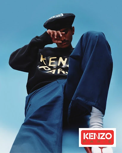 Dark-skinned adult male in a black sweatshirt with the Kenzo Paris logo, blue pants, and a black cap, posing against a blue background.