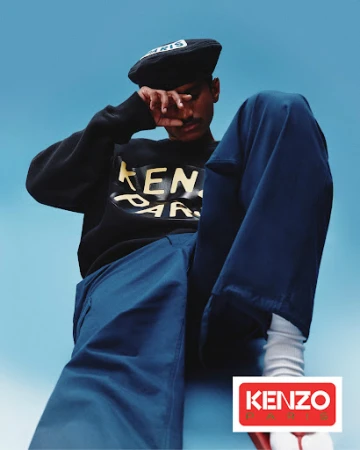 Dark-skinned adult male in a black sweatshirt with the Kenzo Paris logo, blue pants, and a black cap, posing against a blue background.