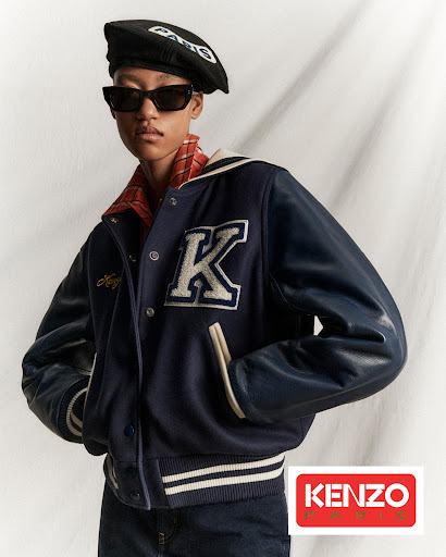 An adult fashion model wearing a beret and sunglasses poses in a letterman jacket against a neutral backdrop, with the Kenzo logo visible.