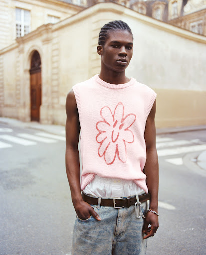 Young Black man in a pink knit vest with a floral design, standing on a city street with light-colored buildings in the background.