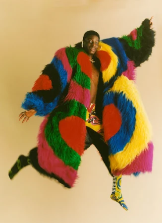 A Black man in a vibrant multi-colored faux fur coat jumping mid-air against a warm neutral studio background.