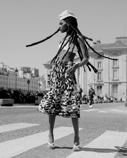 Black and white medium shot of a black female model in a skirt and top on a city crosswalk, hair blowing in wind, Parisian architecture.