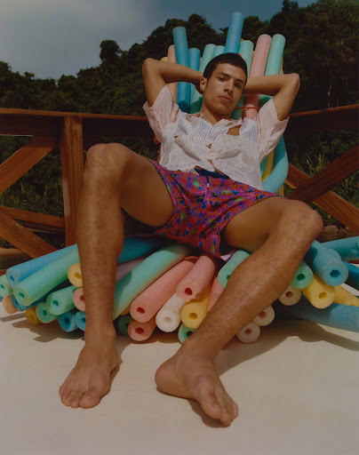 Man in patterned swim trunks lounges on chair made of colorful pool noodles on a sunny deck surrounded by lush greenery.