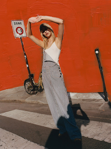 A stylish woman in a jumpsuit poses on a street with a scooter and orange wall backdrop, creating a fashion-forward editorial photograph.