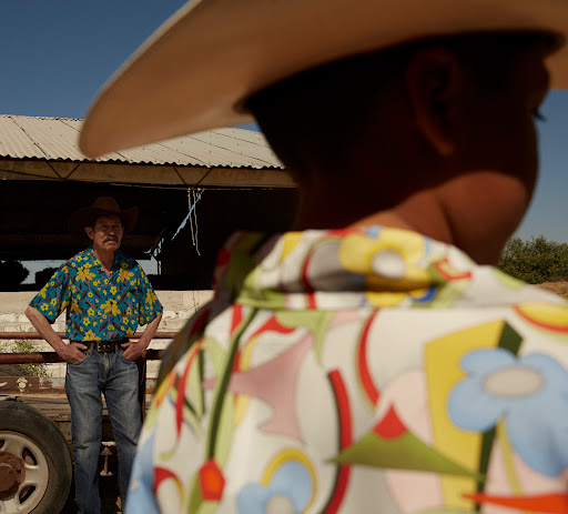 Two men in patterned shirts and cowboy hats stand in a rural setting under a bright blue sky, creating a casual and stylish scene.