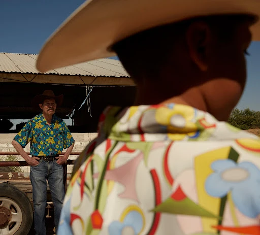 Two men in patterned shirts and cowboy hats stand in a rural setting under a bright blue sky, creating a casual and stylish scene.