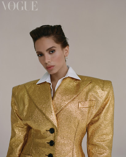 Portrait of woman in a gold, shimmering blazer with a white collared shirt underneath against a neutral background.