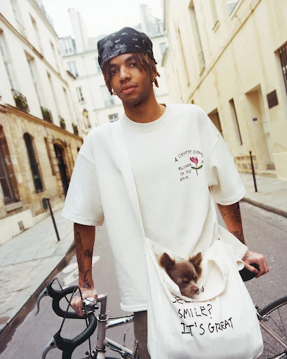 Young man on a bicycle with a small dog in a tote bag, wearing a white t-shirt and a bandana, on a European city street.