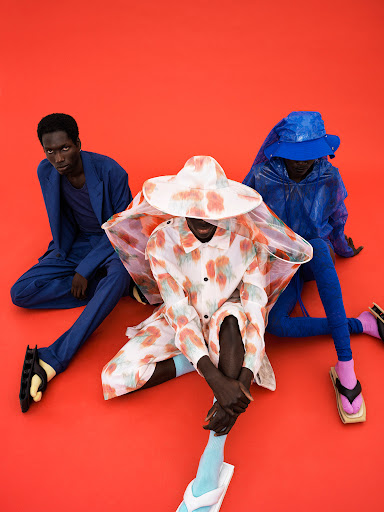 Three dark-skinned adult fashion models in vibrant clothing sit closely together on a red background, showcasing unique textures and styles.