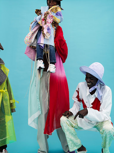 Three individuals in colorful avant-garde fashion, one being held aloft, posing against a vivid turquoise backdrop in an indoor setting.