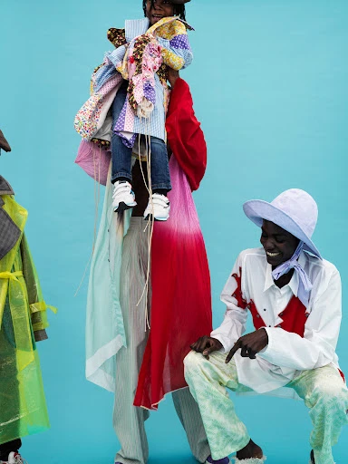 Three individuals in colorful avant-garde fashion, one being held aloft, posing against a vivid turquoise backdrop in an indoor setting.