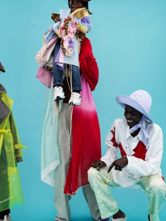 Three individuals in colorful avant-garde fashion, one being held aloft, posing against a vivid turquoise backdrop in an indoor setting.