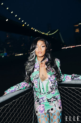 A woman with black and white hair poses in a patterned suit with a bridge and dark sky in the background, seen at night.