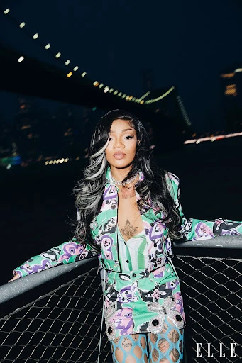 A woman with black and white hair poses in a patterned suit with a bridge and dark sky in the background, seen at night.
