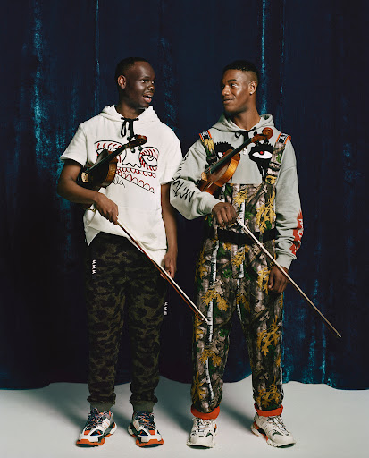 Two men in patterned outfits stand in front of a dark backdrop holding violins, looking to the side in a fashion studio shot.