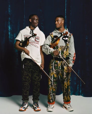 Two men in patterned outfits stand in front of a dark backdrop holding violins, looking to the side in a fashion studio shot.
