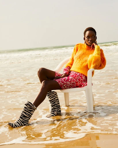 An adult woman sits on a white plastic chair at the beach near the ocean, wearing a bright orange sweater and zebra-print boots.