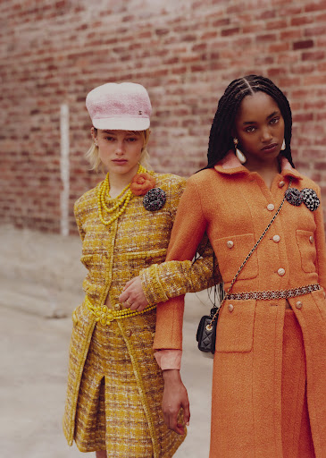 Two fashion models pose outdoors against a brick wall; the model on the left wears a yellow tweed suit and the other model an orange tweed suit.