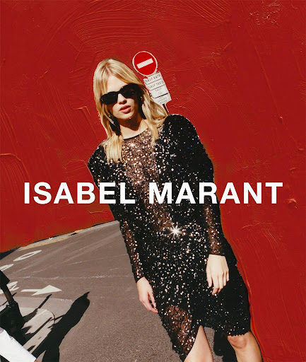 A blonde fashion model in a black sequin dress poses on a city street in front of a red wall with a traffic sign for Isabel Marant.