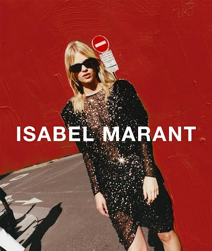 A blonde fashion model in a black sequin dress poses on a city street in front of a red wall with a traffic sign for Isabel Marant.