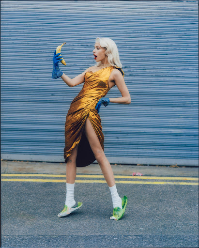Fashionable woman in a gold dress and blue gloves holding a banana, standing on a street with a blue garage door background.