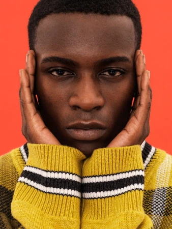 Close-up portrait of a Black man wearing a yellow plaid sweater with hands pressed against his temples against a red backdrop.