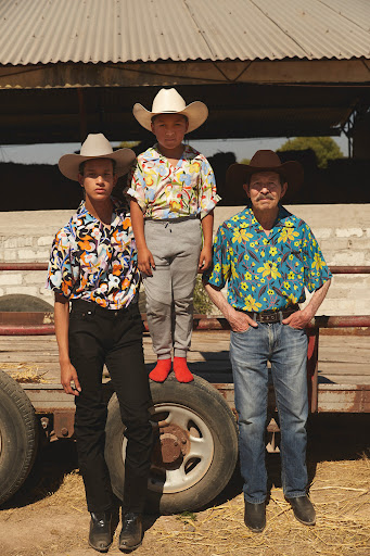 Three people wearing cowboy hats and patterned shirts pose outdoors, one standing on a trailer with large tires, rural, sunny day.