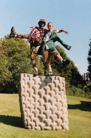 Two adult models pose atop a modern stone sculpture in a park setting, showcasing colorful patterned fashion in an outdoor editorial shot.