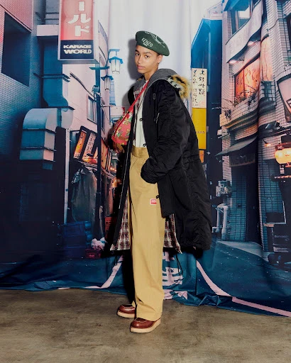 Young man in a black coat, beret, and khaki pants poses against a backdrop of a stylized Japanese street scene with neon signs.