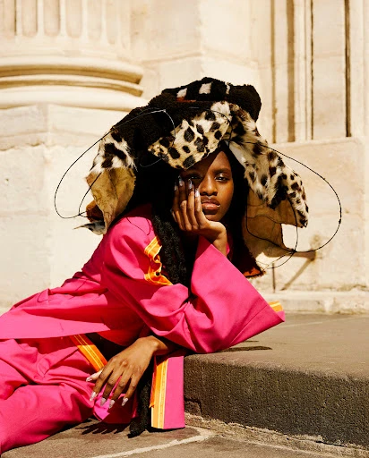 Portrait of an adult female posing on stone steps wearing pink pantsuit and large animal print hat with architectural background.