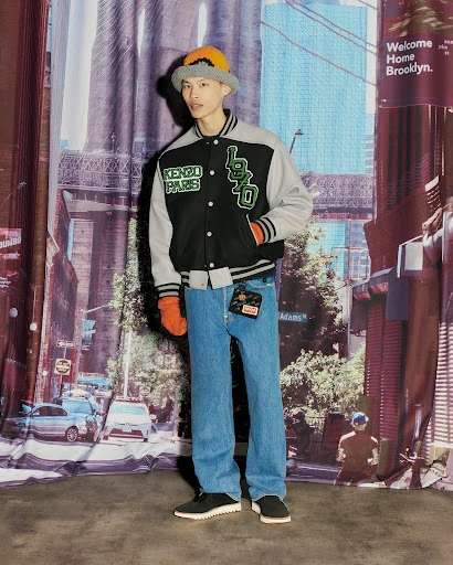 Man in Kenzo jacket stands against a Brooklyn backdrop, wearing jeans, a bucket hat, and orange gloves in a fashion shot.