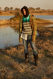 Model wearing a black hoodie under a plaid flannel shirt, patterned sweater, and paint-splattered jeans standing near water.