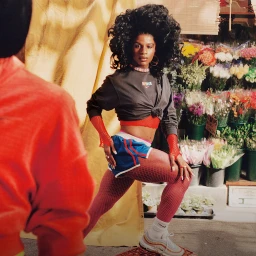 Black woman with voluminous curly hair wearing dark top, red fishnet sleeves, and blue shorts posing near a flower shop