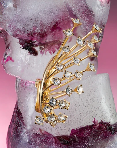 Close-up shot of a gold-tone ear cuff with rhinestones on an ice block with purple flowers. Beauty shot for fashion accessory.