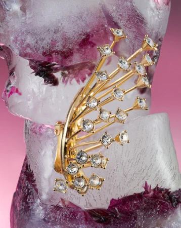 Close-up shot of a gold-tone ear cuff with rhinestones on an ice block with purple flowers. Beauty shot for fashion accessory.