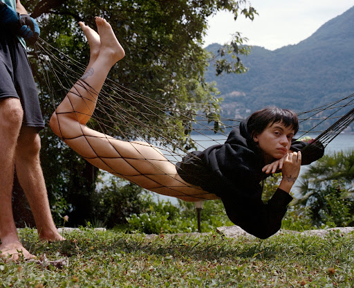 Person resting inside a suspended net hammock above grassy ground with distant mountains across water in the background.