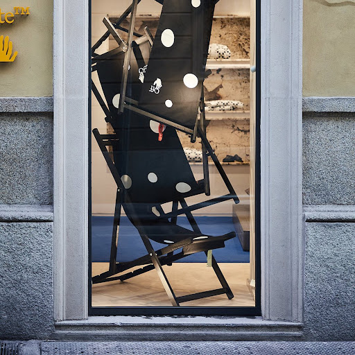 Three black and white polka dot beach chairs stacked inside a storefront window display with a concrete wall and sidewalk outside.