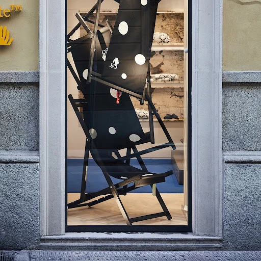 Three black and white polka dot beach chairs stacked inside a storefront window display with a concrete wall and sidewalk outside.