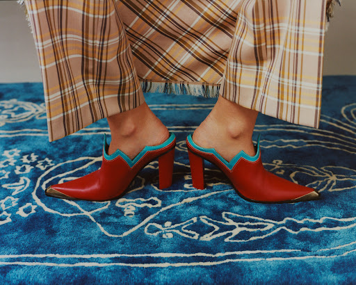 Close-up of a pair of red leather heeled mules with turquoise trim worn with plaid pants on a patterned blue rug.