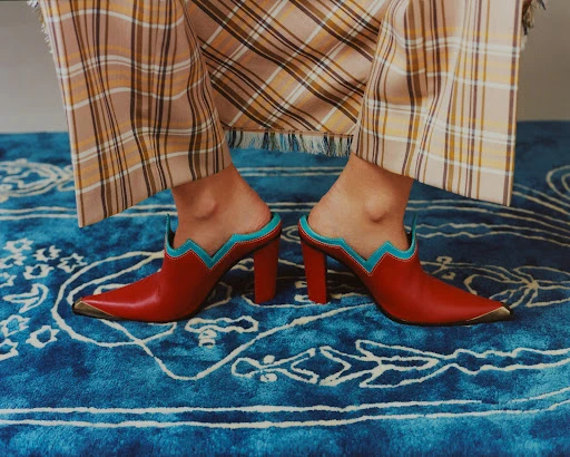 Close-up of a pair of red leather heeled mules with turquoise trim worn with plaid pants on a patterned blue rug.