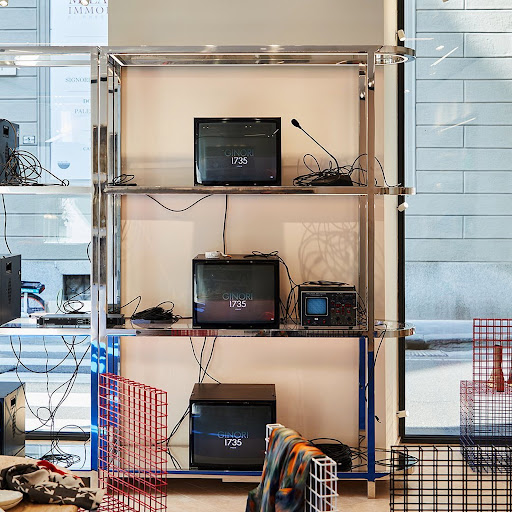 Chrome shelving unit with old televisions displaying 'Ginori 1735,' surrounded by speaker equipment, grid structures, and patterned textiles.