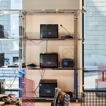 Chrome shelving unit with old televisions displaying 'Ginori 1735,' surrounded by speaker equipment, grid structures, and patterned textiles.