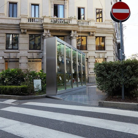 Modern glass bus stop shelter in urban setting against a building facade, with a pedestrian crosswalk in the foreground.