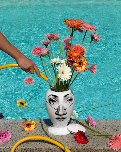 Close-up of a vase with a face design filled with Gerbera daisies being watered near a pool on a sunny day with a yellow hose.