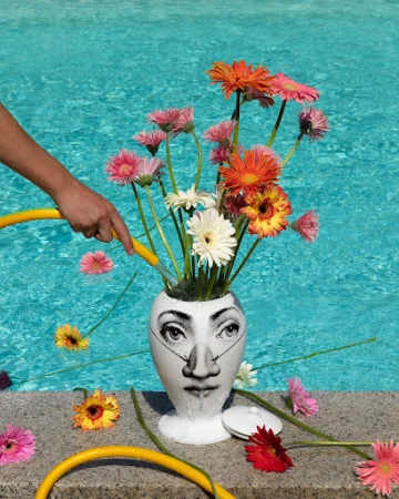 Close-up of a vase with a face design filled with Gerbera daisies being watered near a pool on a sunny day with a yellow hose.