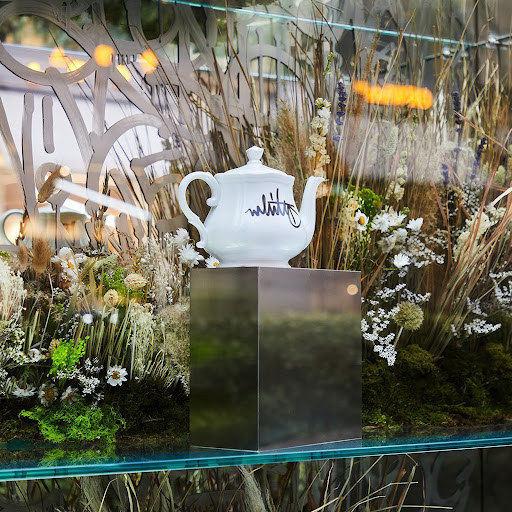 Elegant white teapot on a metallic stand in a window display, surrounded by dried flowers and grasses with a glass pane background.