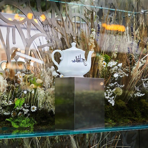 Elegant white teapot on a metallic stand in a window display, surrounded by dried flowers and grasses with a glass pane background.