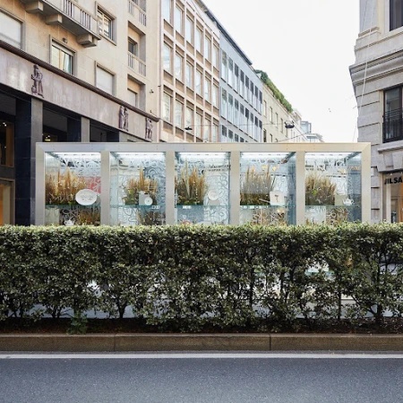 Urban retail storefront display with floral arrangements behind glass panels, framed by modern architecture in an Italian city street.