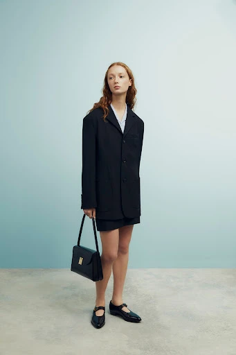 A woman in a dark blazer and skirt, holding a purse, stands in a studio with a light blue backdrop and neutral flooring.