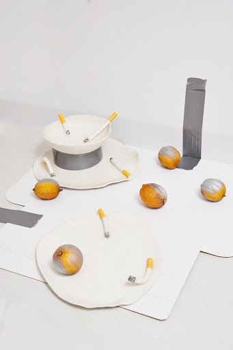 Still life of white ceramic dishes arranged with orange fruit and white cigarettes in a minimalist, modern art photograph.